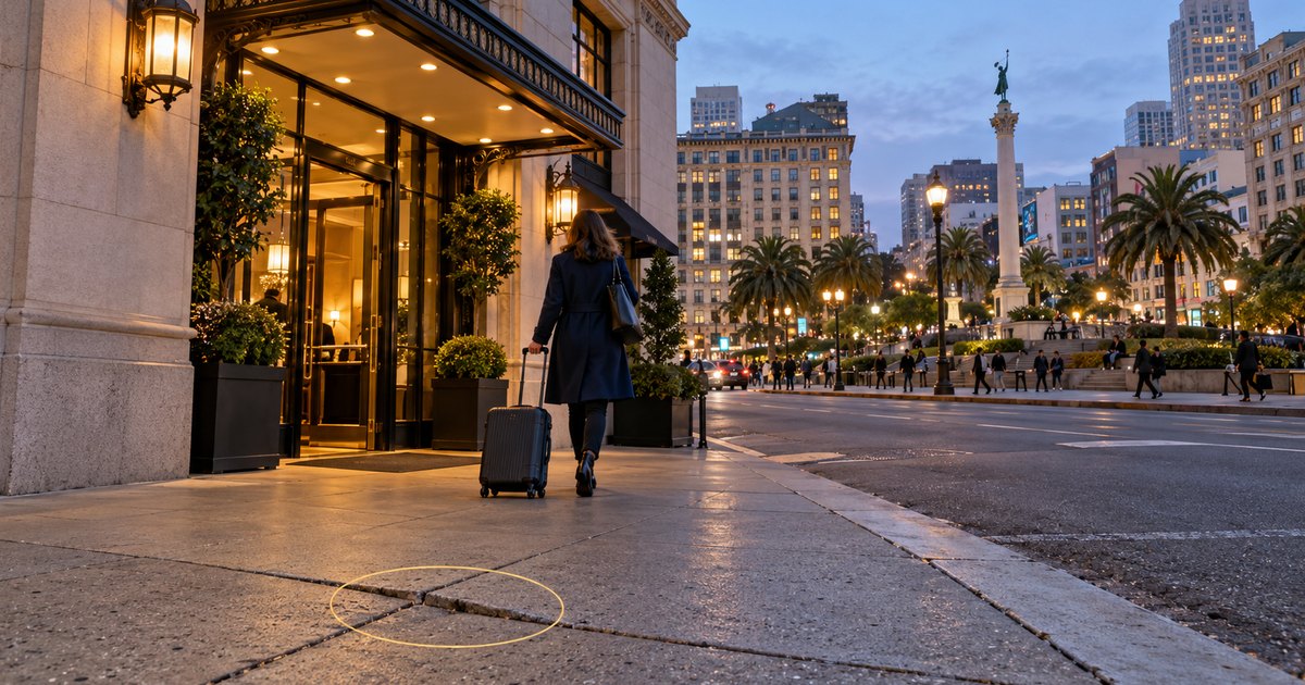 A guest with a rolling suitcase walks toward a hotel entrance on Union Square in San Francisco at dusk, with a gold measurement ring around a cracked sidewalk joint in the foreground